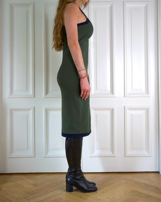 Woman wearing a moss green dress and black boots standing against a white paneled wall.