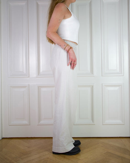 Woman wearing a fitted white tank top with beige striped wide pants and black boots, standing against a white paneled wall.