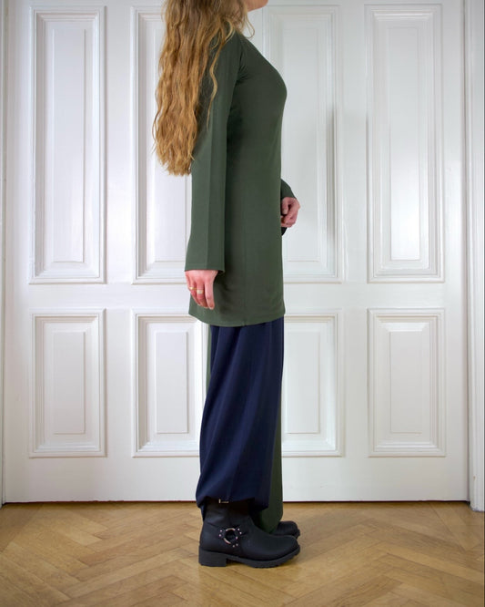 Woman wearing a green dress with long wide sleeves and blue and green pants standing against a white paneled wall.