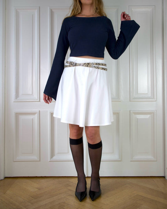 Woman wearing a navy top with long wide sleeves and white a-lined skirt with a snake patterned belt against a white paneled wall.