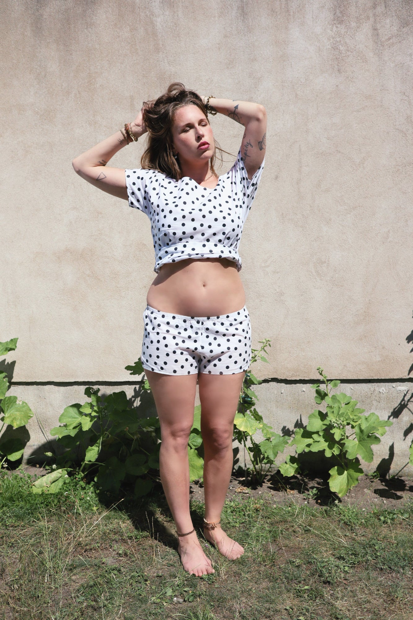 A woman wearing a matching white short-sleeve crop top and shorts set with black polka dots, standing outdoors against a beige stucco wall with green plants at her feet.