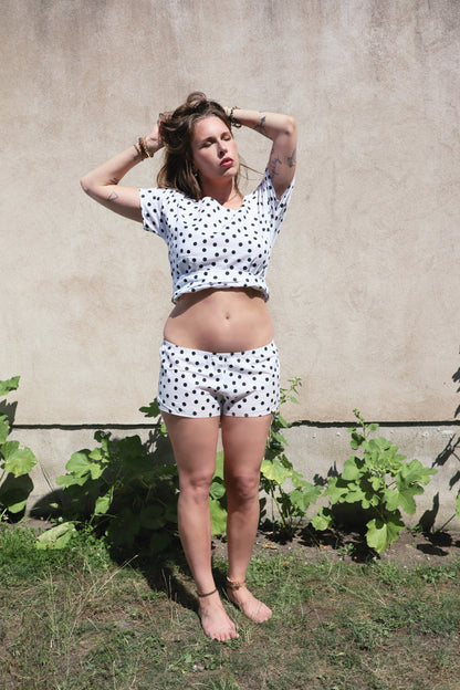 A woman wearing a matching white short-sleeve crop top and shorts set with black polka dots, standing outdoors against a beige stucco wall with green plants at her feet.