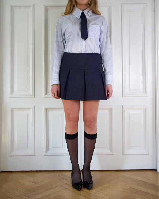 Woman wearing a blue striped collar shirt with a navy blue tie, navy pleated skirt, black stockings, and black shoes against a white paneled wall.