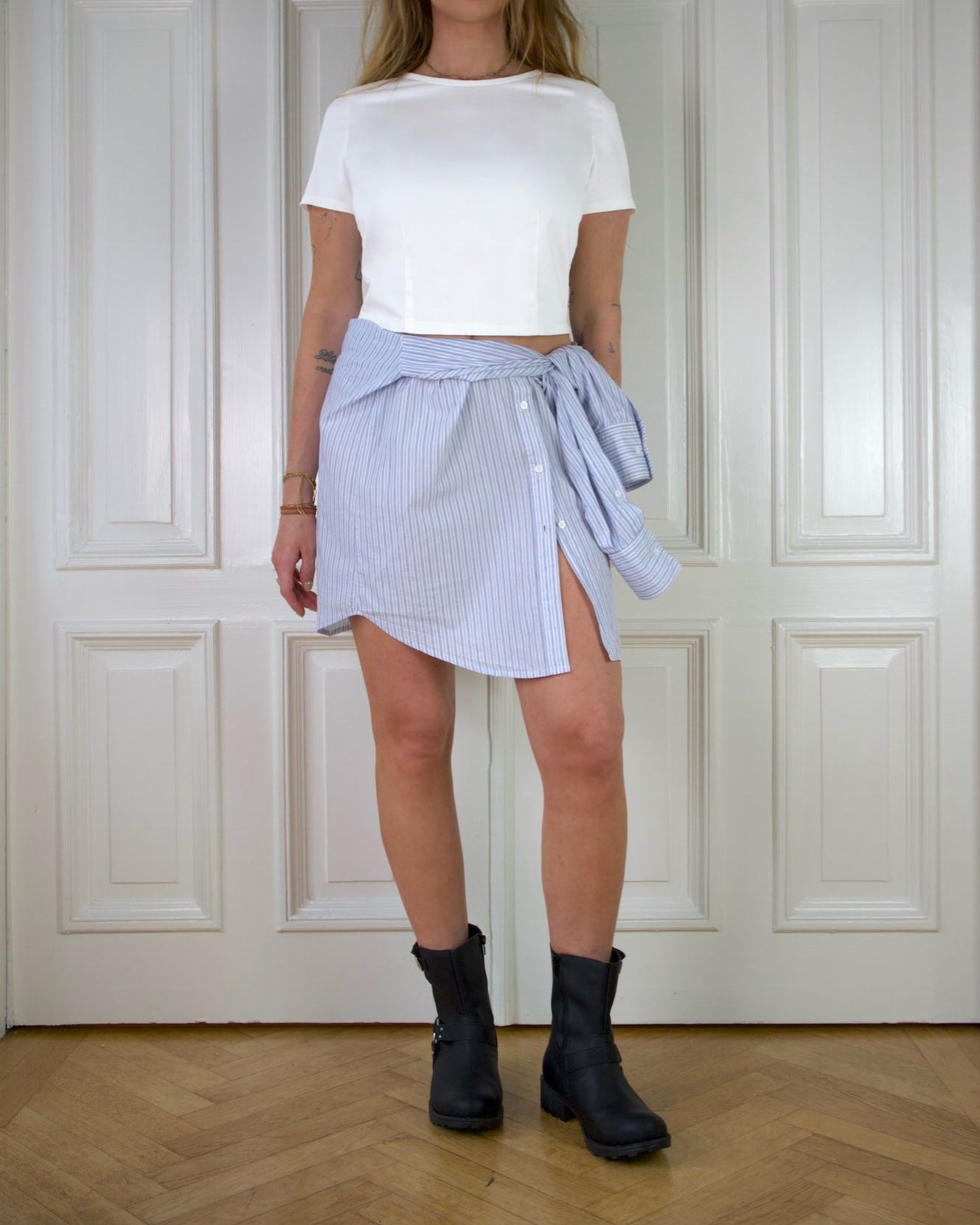 Woman wearing a white fitted top and light blue striped skirt together with black boots against a white paneled wall.