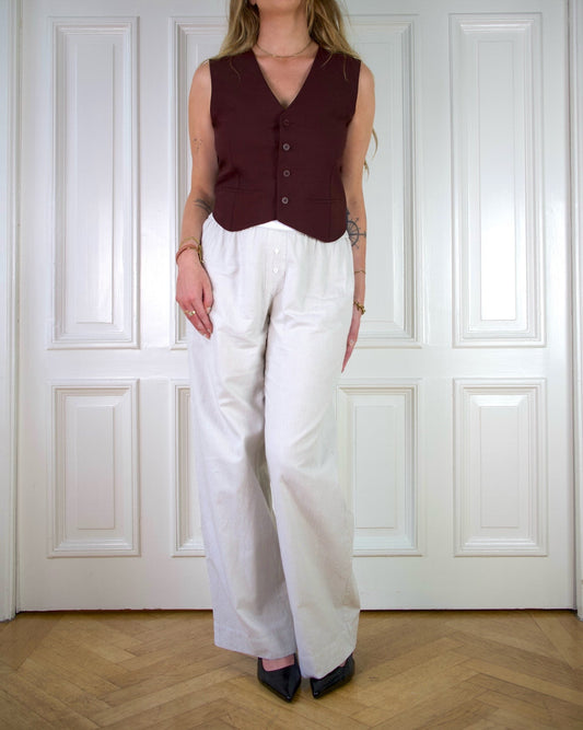 Woman wearing a burgundy vest and beige striped pants standing in front of a white paneled wall.