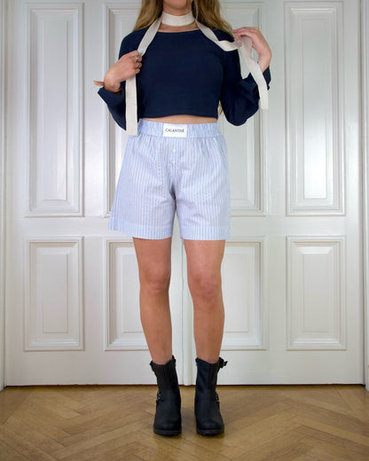 Woman wearing a dark blue top with wide sleeves, light blue striped boxer shorts, and black boots in front of a white door with paneling.