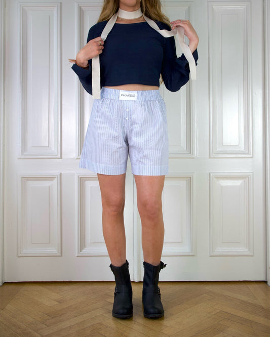 Woman wearing a dark blue top with wide sleeves, light blue striped boxer shorts, and black boots in front of a white door with paneling.