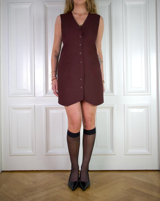 Woman wearing a sleeveless burgundy long vest dress with black stockings and slingback heels, against a white paneled wall.