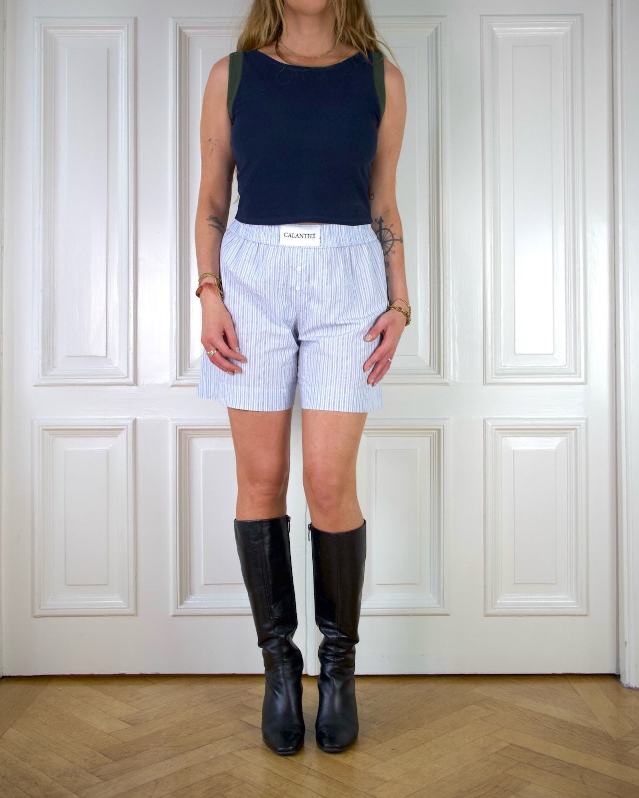 Woman wearing a navy tank top with green edges and blue striped boxer shorts standing in front of a white paneled wall.