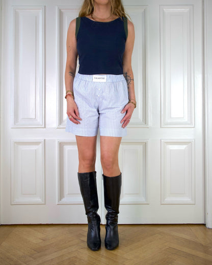 Woman wearing a navy tank top with green edges and blue striped boxer shorts standing in front of a white paneled wall.