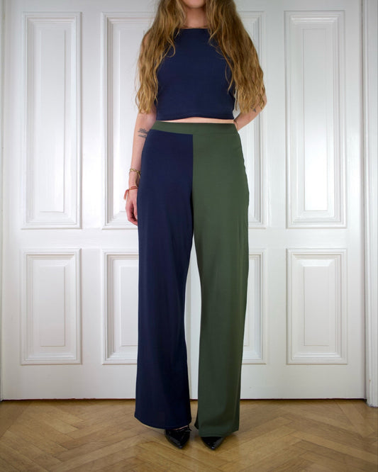Person wearing a blue top and green nad navy blue pants standing in front of a white paneled wall.