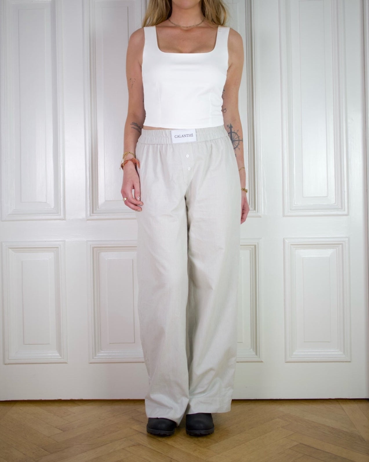 Woman wearing a white fitted tank top and beige striped long pants standing against a white paneled wall.