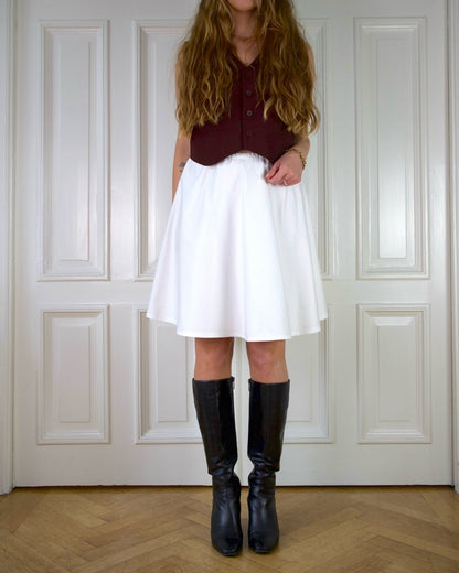 Woman wearing a white skirt, burgundy vest, and black boots standing in front of a white paneled wall.