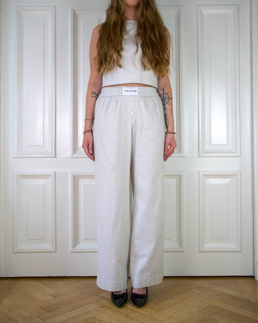 Woman wearing a beige striped sleeveless top and wide-leg pants standing against a white paneled wall.