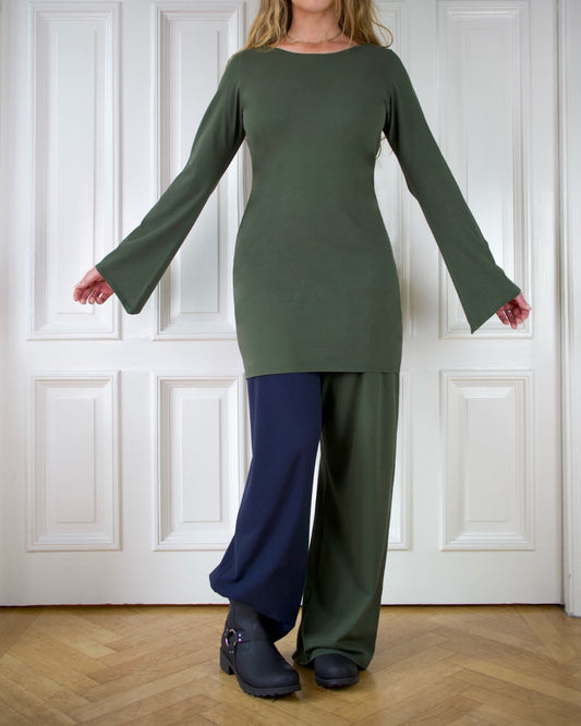 Woman wearing a long green dress with long wide sleeves, paired with blue and green pants against a white paneled wall.