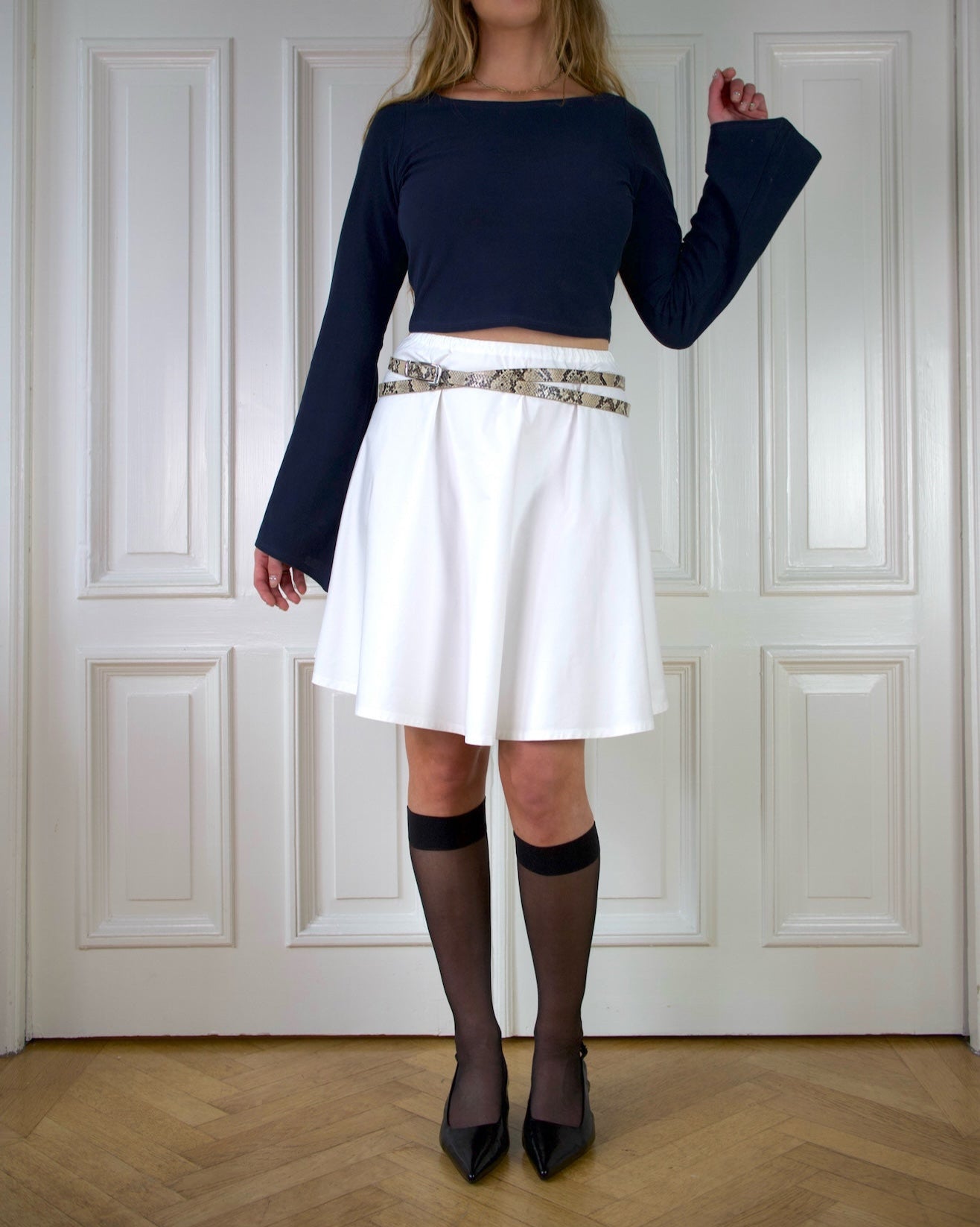 Woman wearing a navy top with long wide sleeves and white a-lined skirt with a snake patterned belt against a white paneled wall.