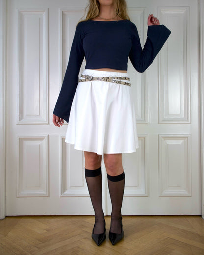 Woman wearing a navy top with long wide sleeves and white a-lined skirt with a snake patterned belt against a white paneled wall.