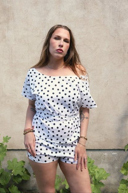 A woman modeling a white off-the-shoulder top with black polka dots, standing against a textured concrete wall with green plants at the bottom.